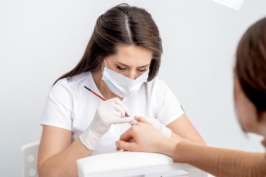 Manicure master with protective mask applying transparent nail polish on female nails in beauty salon