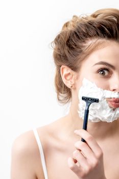 Beautiful young caucasian woman shaving her face by razor on white background. Pretty woman with shaving foam on her face