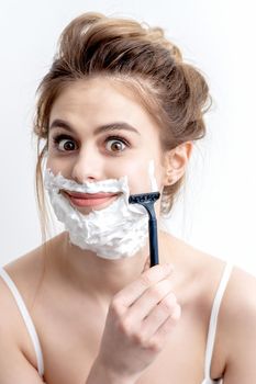 Beautiful young caucasian woman shaving her face by razor on white background. Pretty woman with shaving foam on her face