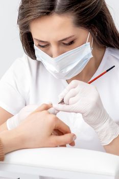 Manicure master with protective mask applying transparent nail polish on female nails in beauty salon