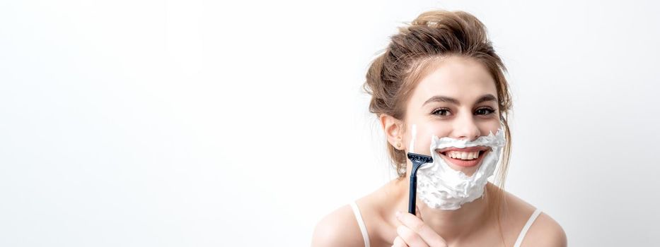 Beautiful young caucasian woman shaving her face by razor on white background. Pretty woman with shaving foam on her face