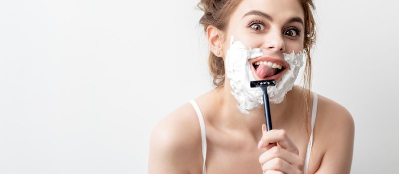 Beautiful young caucasian woman shaving her face by razor on white background. Pretty woman with shaving foam on her face