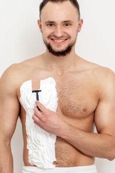 Young caucasian man with beard holds razor shaves his chest with white shaving foam on white background. Man shaving his torso