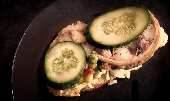 On a glass plate is a sandwich with a meat snack and vegetables. Top view, dark background, copy space