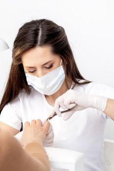 Manicurist in protective mask using manicure cutter to remove cuticle of female nails in manicure salon