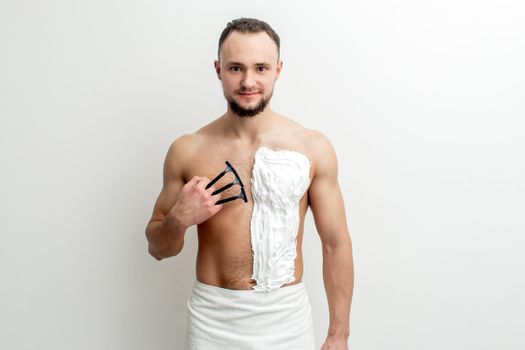 Young caucasian man with beard holds razor shaves his chest with white shaving foam on white background. Man shaving his torso