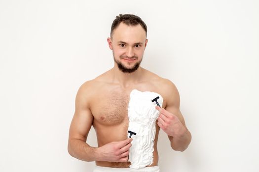 Young caucasian man with beard holds razor shaves his chest with white shaving foam on white background. Man shaving his torso