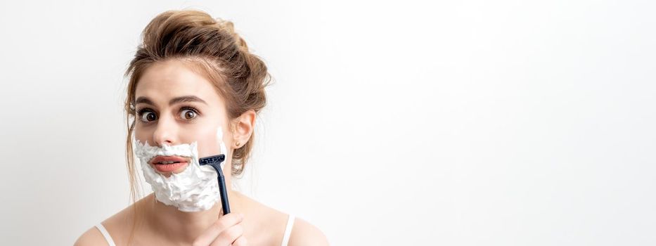 Beautiful young caucasian woman shaving her face by razor on white background. Pretty woman with shaving foam on her face