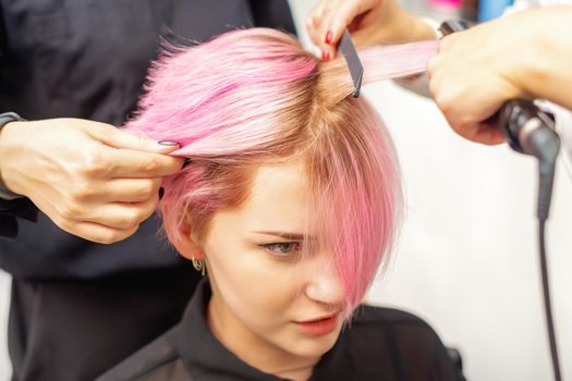Hairdresser straights pink hair of young woman by hair iron in hairdress salon