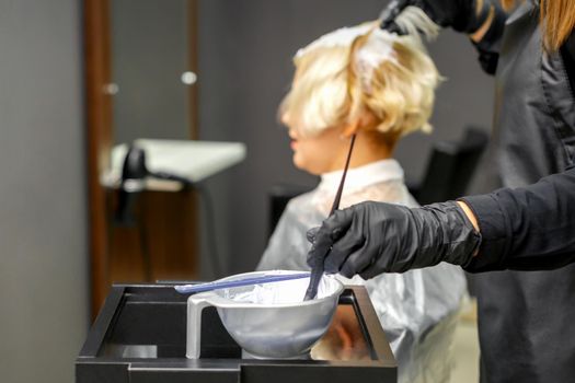 Hairdresser in black gloves dyes hair of young woman in white color in hairdress salon