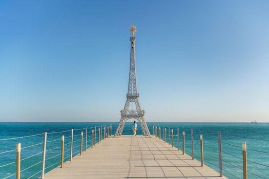 Large model of the Eiffel Tower on the beach. A woman walks along the pier towards the tower, wearing a blue jacket and white jeans