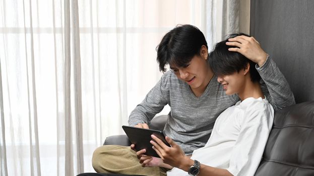 Romantic same sex male couple using digital tablet together on sofa, spending time together at home. Homosexual relationships concept.