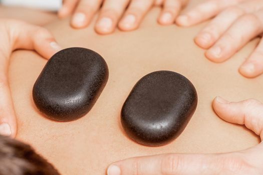 Hands of massage therapists doing back massage while hot stones on back of man close up in spa.