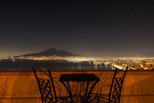 Gulf of Naples evening cityscape with scene of the active volcano, Mount Vesuvius, visible above a table and two chairs from a terrace in Pompeii. City lights illuminate the the night sky.