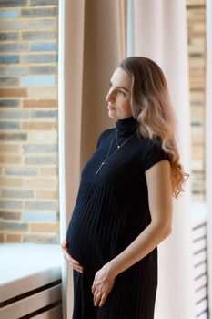 Thoughtful pregnant woman caressing her belly and looking out the window.