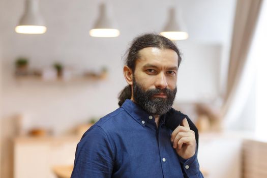 Portrait of a cute long-haired man with a beard in a blue shirt and jeans against the background of a bright, warm room