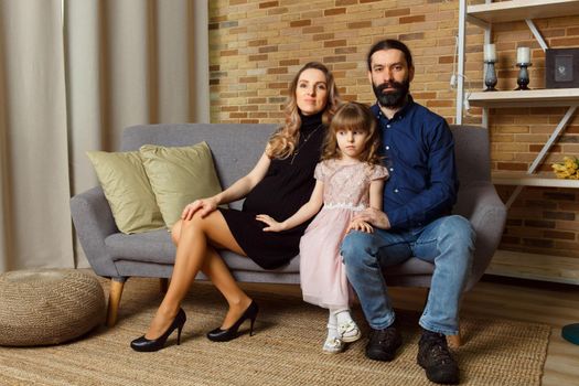 Happy young father, mother and daughter sit on wicker sofa at home. The image of a happy family expecting the second child, studio