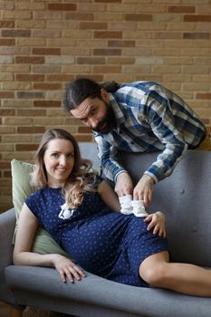A man and his pregnant wife pose on a sofa in their home. They are holding baby booties that will be worn by their future child. The concept of waiting for a child.