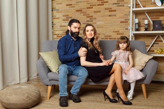 Happy young father, mother and daughter sit on wicker sofa at home. The image of a happy family expecting the second child, studio