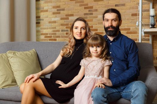 Happy young father, mother and daughter sit on wicker sofa at home. The image of a happy family expecting the second child, studio