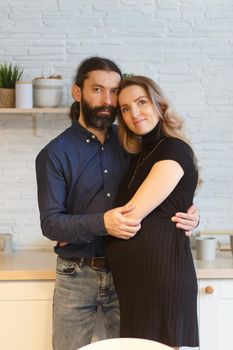 Man embracing pregnant partner in kitchen. Adult family pregnancy concept. Future parents in home outfit embrace standing in the kitchen, looking at each other. Healthy Lifestyle.