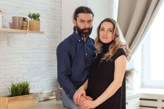 Man embracing pregnant partner in kitchen. Adult family pregnancy concept. Future parents in home outfit embrace standing in the kitchen, looking at each other, kissing. Healthy Lifestyle.