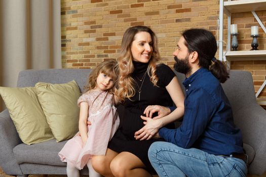 Happy young father, mother and daughter sit on wicker sofa at home. The image of a happy family expecting the second child, studio