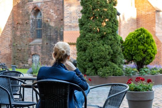 Young blonde woman sits on wicker chair in street cafe and looks at historic red brick building. Selective focus. View from the back.