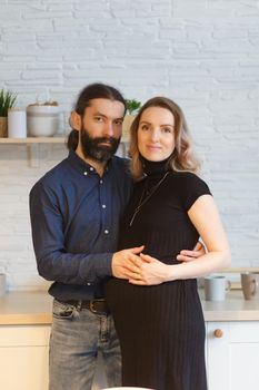 Man embracing pregnant partner in kitchen. Adult family pregnancy concept. Future parents in home outfit embrace standing in the kitchen, looking at each other. Healthy Lifestyle.