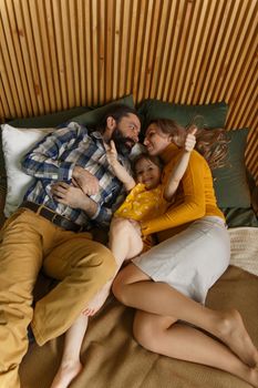 Happy family is having fun in bedroom. Enjoying being together. Parents are tickling their little daughter while lying in bed. Father, daughter and pregnant mother.