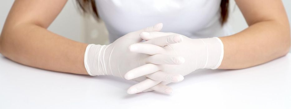 Hands in protective white medical gloves of the woman sitting at the white table. Crossed fingers in gloves