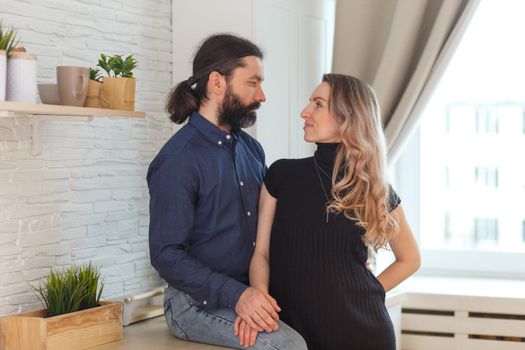 Man embracing pregnant partner in kitchen. Adult family pregnancy concept. Future parents in home outfit embrace standing in the kitchen, looking at each other, kissing. Healthy Lifestyle.