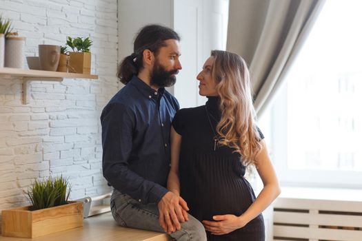 Man embracing pregnant partner in kitchen. Adult family pregnancy concept. Future parents in home outfit embrace standing in the kitchen, looking at each other, kissing. Healthy Lifestyle.