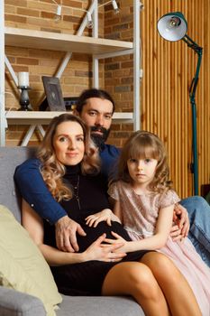 Happy young father, mother and daughter sit on wicker sofa at home. The image of a happy family expecting the second child, studio