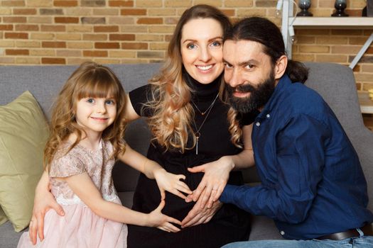 Happy young father, mother and daughter sit on wicker sofa at home. The image of a happy family expecting the second child, studio