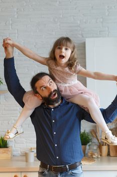 Happy little daughter sitting on father neck piggybacking playing active game at home. Smiling dad holding cute girl with arms outstretched carrying on back. Family spending weekend together.