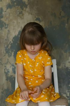 Portrait of a little upset girl in a yellow dress sitting on a chair on an abstract blue background.