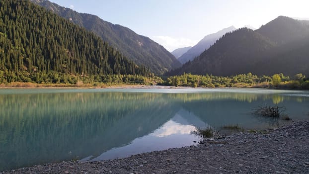 View of the mountain lake Issyk from a height. Turquoise-emerald water from the glacier. Reflection of mountains, forests and the sun on the water. The river flows into the lake. Issyk Dam. Kazakhstan