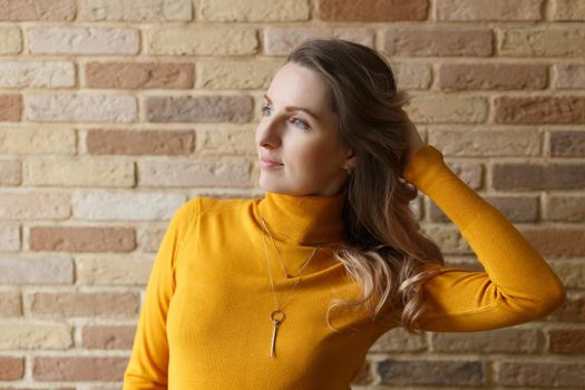 Thoughtful pregnant woman in yellow cardigan caressing her belly and posing near by brick wall