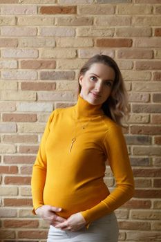 Thoughtful pregnant woman in yellow cardigan caressing her belly and posing near by brick wall