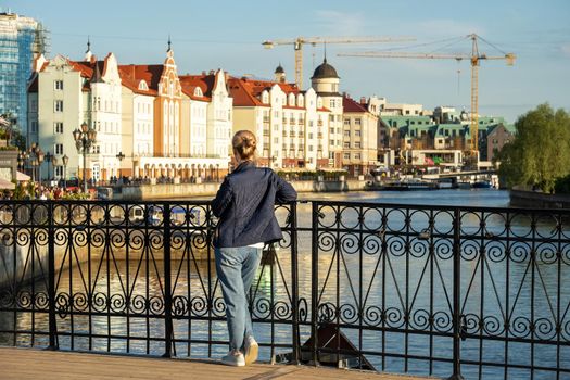 Young blonde woman stands on bridge and looks at beautiful city embankment on spring evening. View from the back. Selective focus.