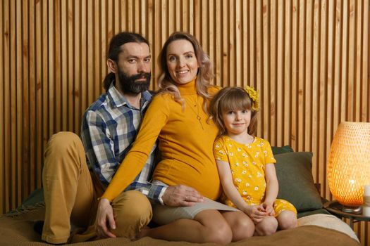 Happy family is having fun in bedroom. Enjoying being together. Parents are tickling their little daughter while lying in bed. Father, daughter and pregnant mother.