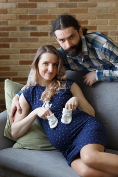 A man and his pregnant wife pose on a sofa in their home. They are holding baby booties that will be worn by their future child. The concept of waiting for a child.