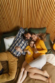 Happy family is having fun in bedroom. Enjoying being together. Parents are tickling their little daughter while lying in bed. Father, daughter and pregnant mother.