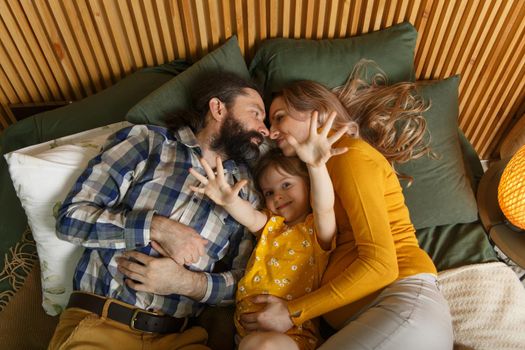 Happy family is having fun in bedroom. Enjoying being together. Parents are tickling their little daughter while lying in bed. Father, daughter and pregnant mother.