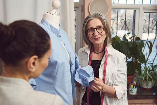 Skilled senior fashion designer takes measurement and works on improvement details of a blue shirt from new collection on a mannequin dummy in a fashion design and tailoring workshop