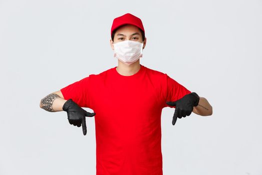 Look here. Serious, confident asian delivery guy with tattooed arm, pointing fingers down to show advertisement promo in protective gloves and medical mask. Courier wearing uniform, delivery parcels.