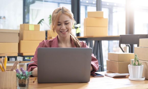 Young beautiful happy asian business woman owner of SME online using laptop receive order from customer with parcel box packaging at her startup home office, online business seller and delivery.