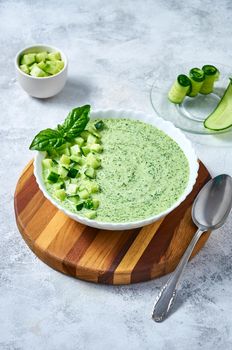 Cucumber Gazpacho - cold summer soup with basil in white bowl on wooden board on light background.