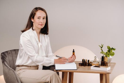 The aromatherapist girl is sitting in her office and holding a bottle of aromatic oil in her hands and writing something down. there are essential oils on the table.